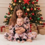 Older sister holding a baby brother, both wearing matching seamless French Bulldog pattern Christmas pajamas, sitting on a white rug in front of a brightly decorated Christmas tree.